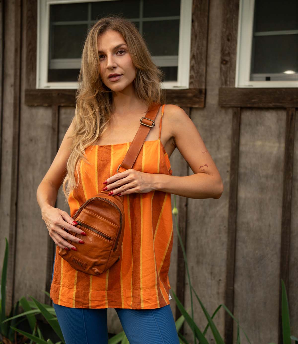 
                  
                    A woman with long blonde hair stands before a wooden building, wearing Roan's Trickle orange striped top, blue pants, and carrying a compact leather bag.
                  
                