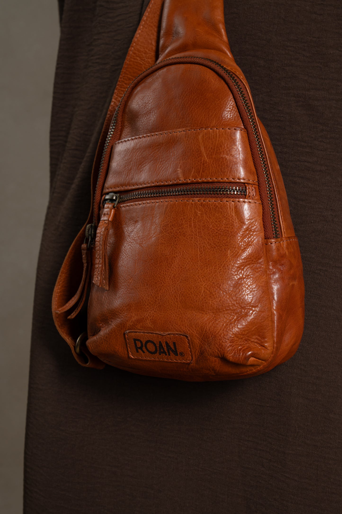 
                  
                    A close-up of the compact Trickle leather bag by Roan, featuring the brand name on a patch, worn over a dark brown garment and highlighting its everyday versatility in a classic crossbody design.
                  
                