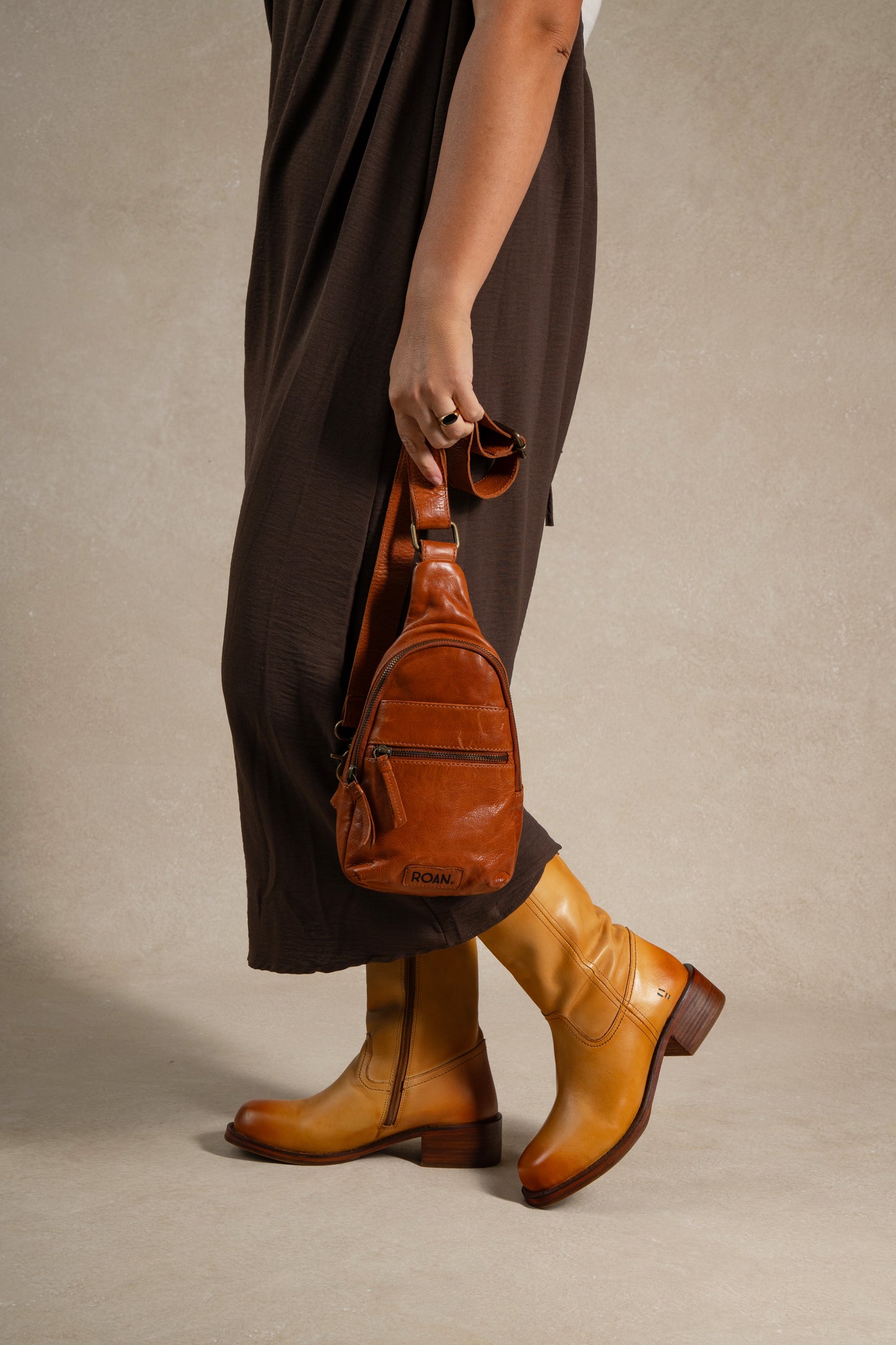 
                  
                    A person in a brown dress and tan boots holds the Roan Trickle compact leather bag, standing against a neutral background to showcase its effortless everyday versatility.
                  
                