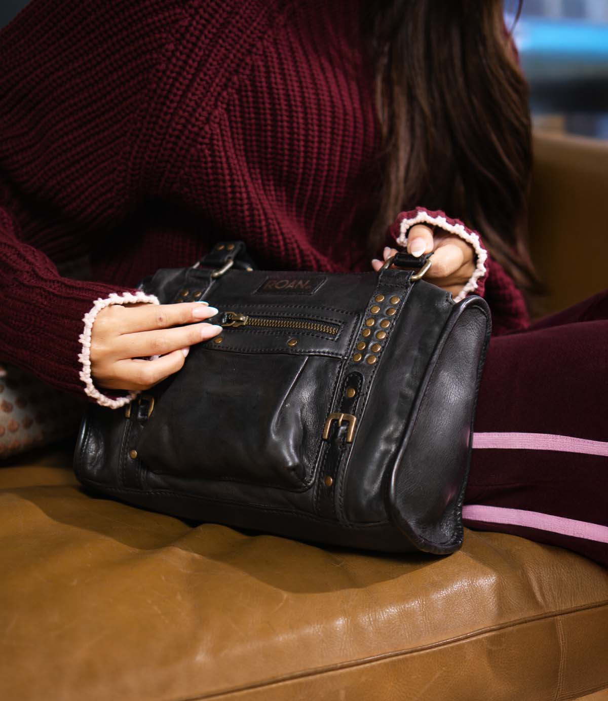 A person holds a structured Roan Visual leather handbag with a front zippered pocket and brass studded accents.