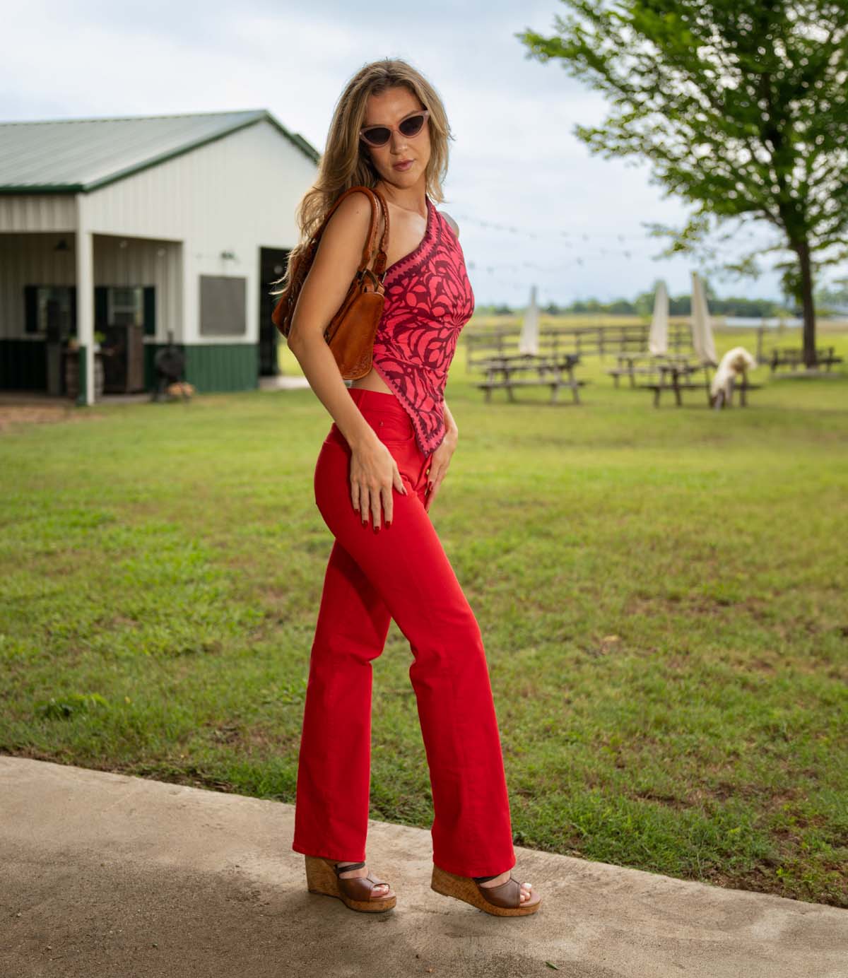 
                  
                    A woman in red pants, a red patterned top, and Roan Decorum leather wedge sandals with a cushioned footbed stands on a covered walkway near grass, picnic tables, and a barn.
                  
                