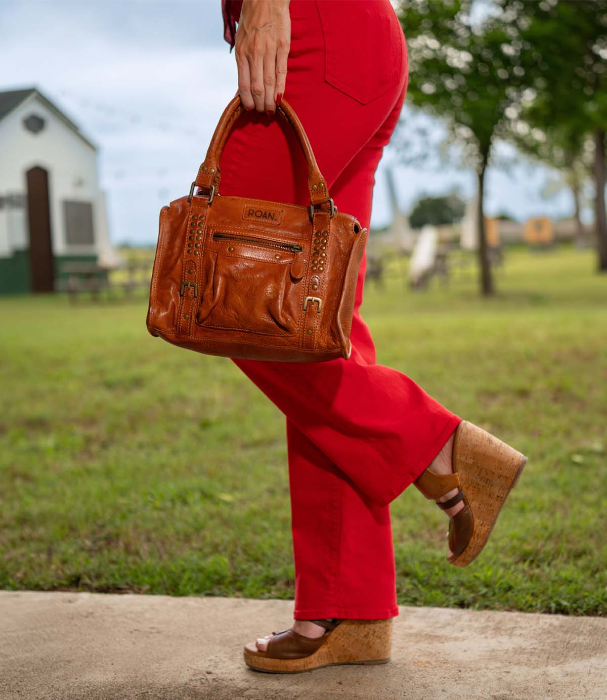 A person in red pants and brown wedge sandals carries the Roan Visual everyday bag with studded accents outdoors on a concrete path, with grass and trees in the background.