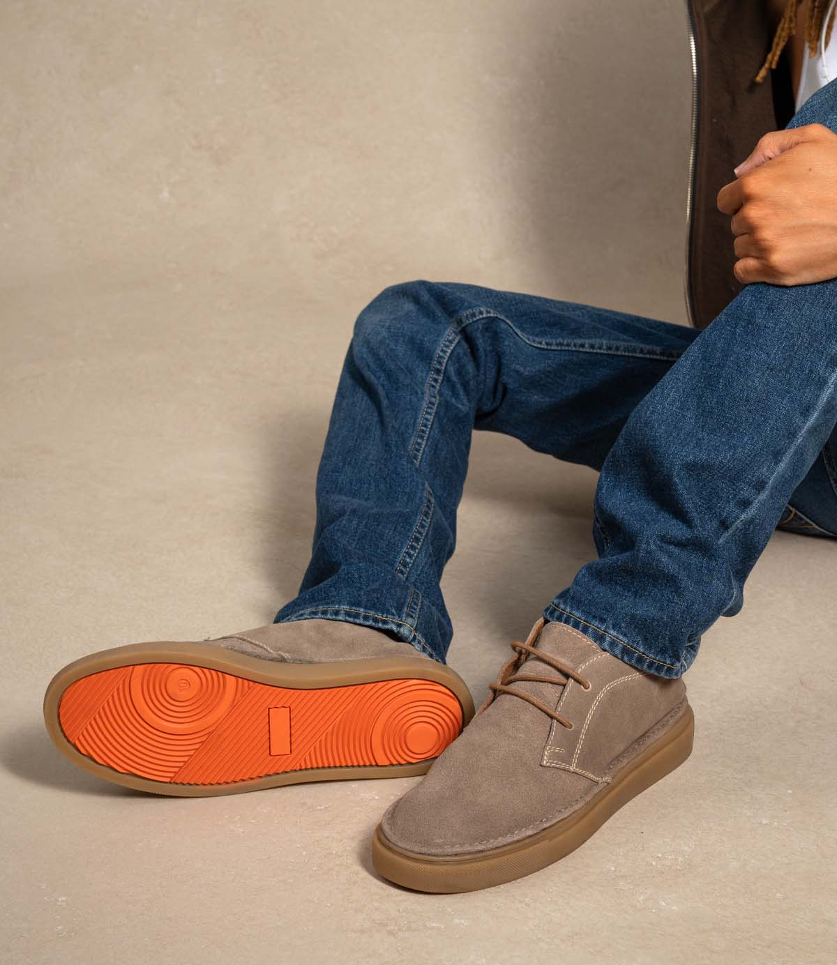
                  
                    A person sits on the floor wearing blue jeans and Roan’s Volunteer soft suede chukka shoes with bright orange, sneaker-style soles; only their legs and one arm are visible.
                  
                