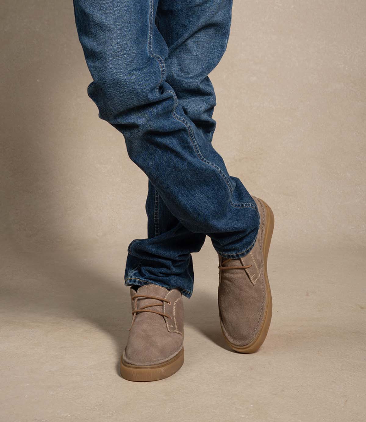 
                  
                    A person wearing blue jeans and the Roan Volunteer beige soft suede chukka shoes with a sneaker-style sole, standing with legs crossed in front of a neutral background.
                  
                