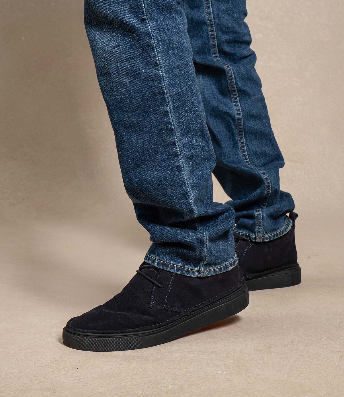 
                  
                    A person wearing blue jeans and Roan’s Volunteer black soft suede chukka shoes with a sneaker-style sole stands on a light-colored floor, feet angled slightly to the side.
                  
                