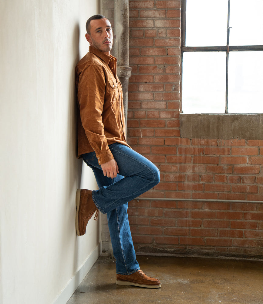 
                  
                    A man in a brown shirt and blue jeans, wearing the Roan Warden chukka boot with a cushioned insole, leans against a white wall with one foot up in an industrial-style room featuring brick walls and a large window.
                  
                