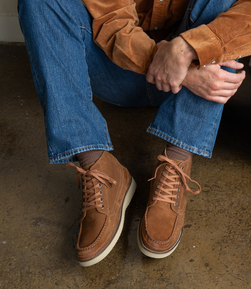 Wearing Roan's Warden moc toe chukka boots with cushioned insoles, blue jeans, and a brown suede shirt, a person sits on a concrete floor with hands resting on one knee.