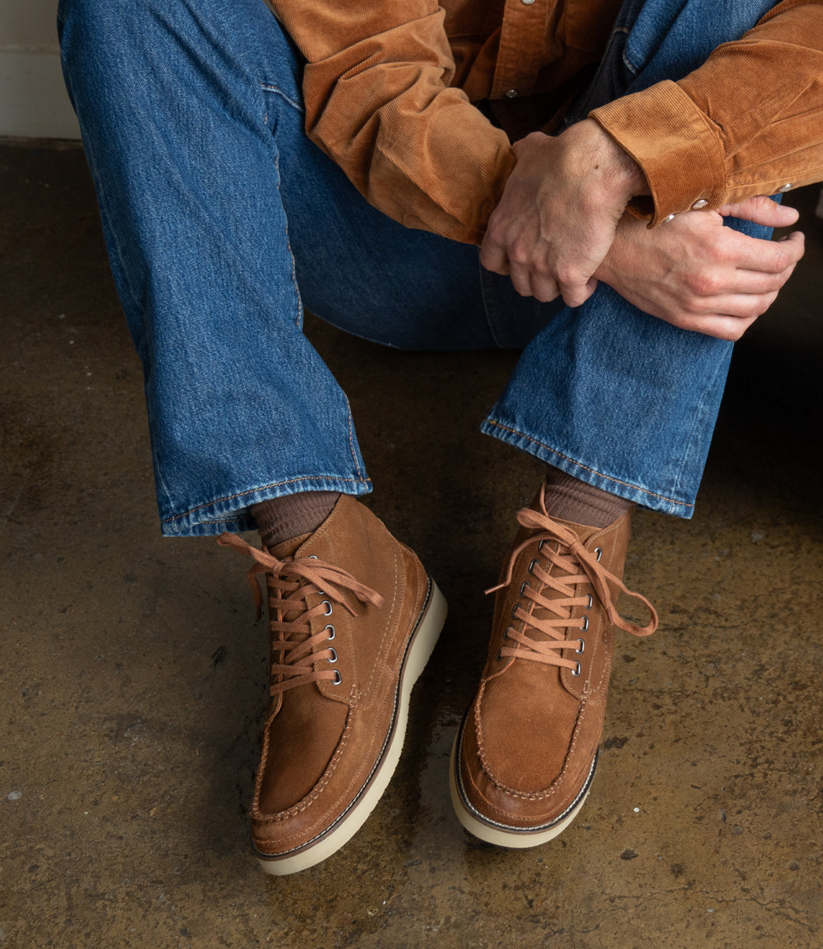 Wearing Roan's Warden moc toe chukka boots with cushioned insoles, blue jeans, and a brown suede shirt, a person sits on a concrete floor with hands resting on one knee.