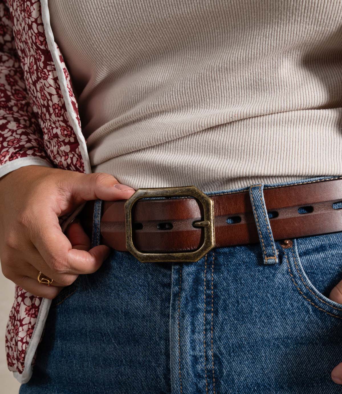 
                  
                    A person wearing a cream ribbed shirt, red patterned jacket, blue jeans, and the Roan “Waymark” brown leather belt—featuring stitched edges and a large square buckle—holding the belt with one hand.
                  
                