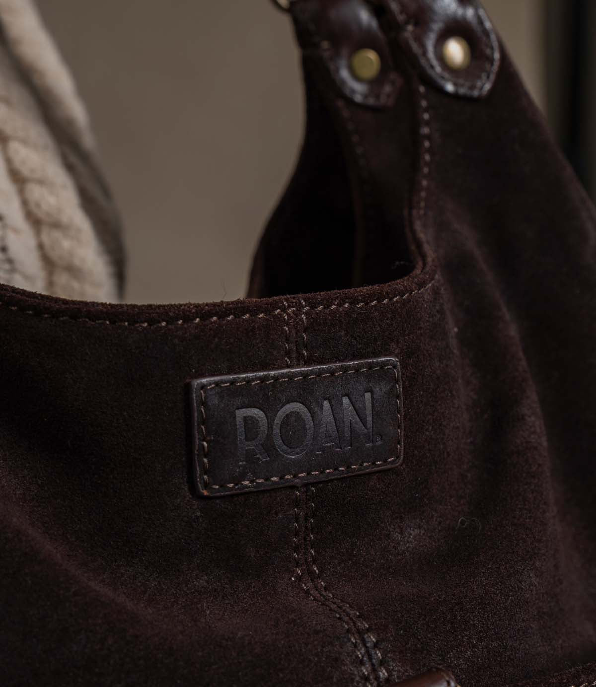 
                  
                    Close-up of the Winery boot by Roan, featuring dark brown suede with visible stitching, a branded leather patch labeled “Roan,” and a glimpse of shearling lining in the background. The design is enhanced by adjustable buckle straps that offer both style and functionality.
                  
                