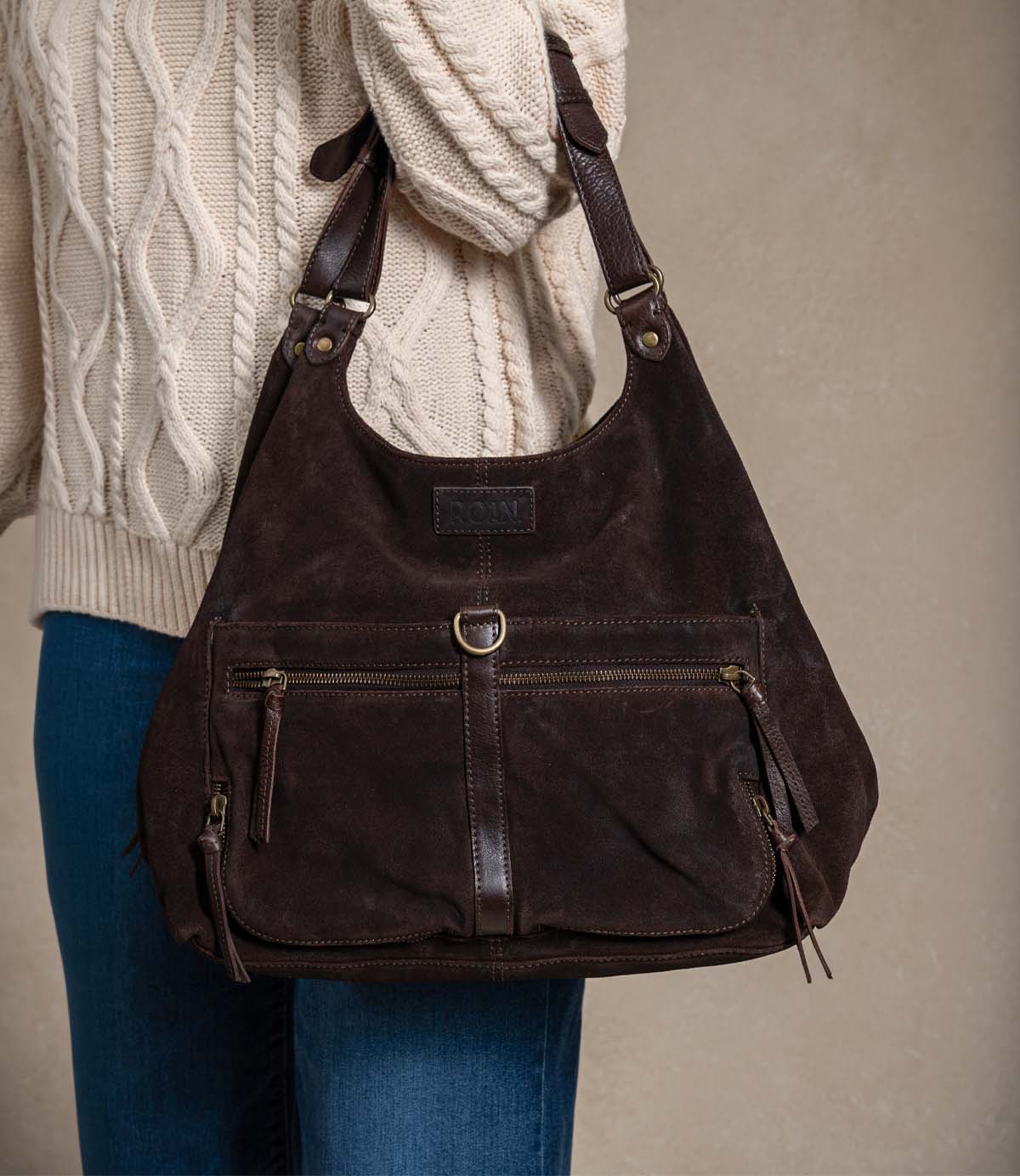 
                  
                    A person wearing a cream cable-knit sweater and blue jeans is holding the Roan Winery—a large dark brown suede shoulder bag with front zippers, leather trim, and expandable zip pockets.
                  
                