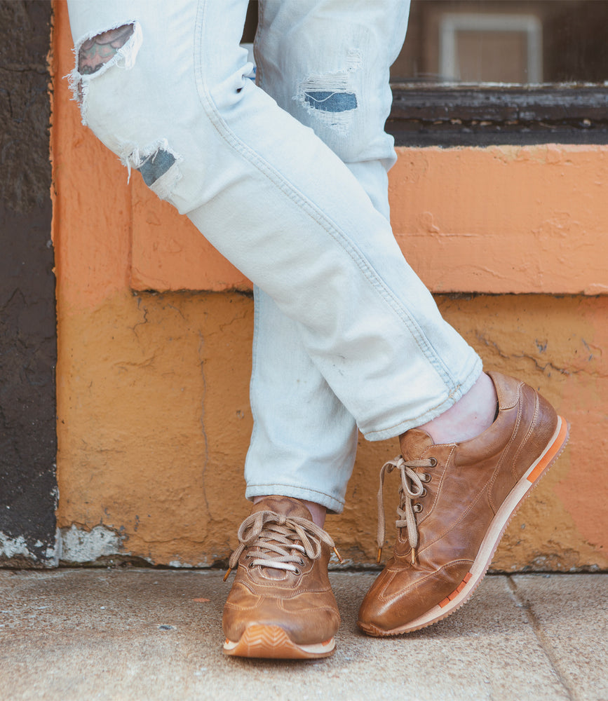 Person wearing ripped jeans and Roan Yann brown high-quality leather low profile sneakers leaning against an orange and yellow wall.