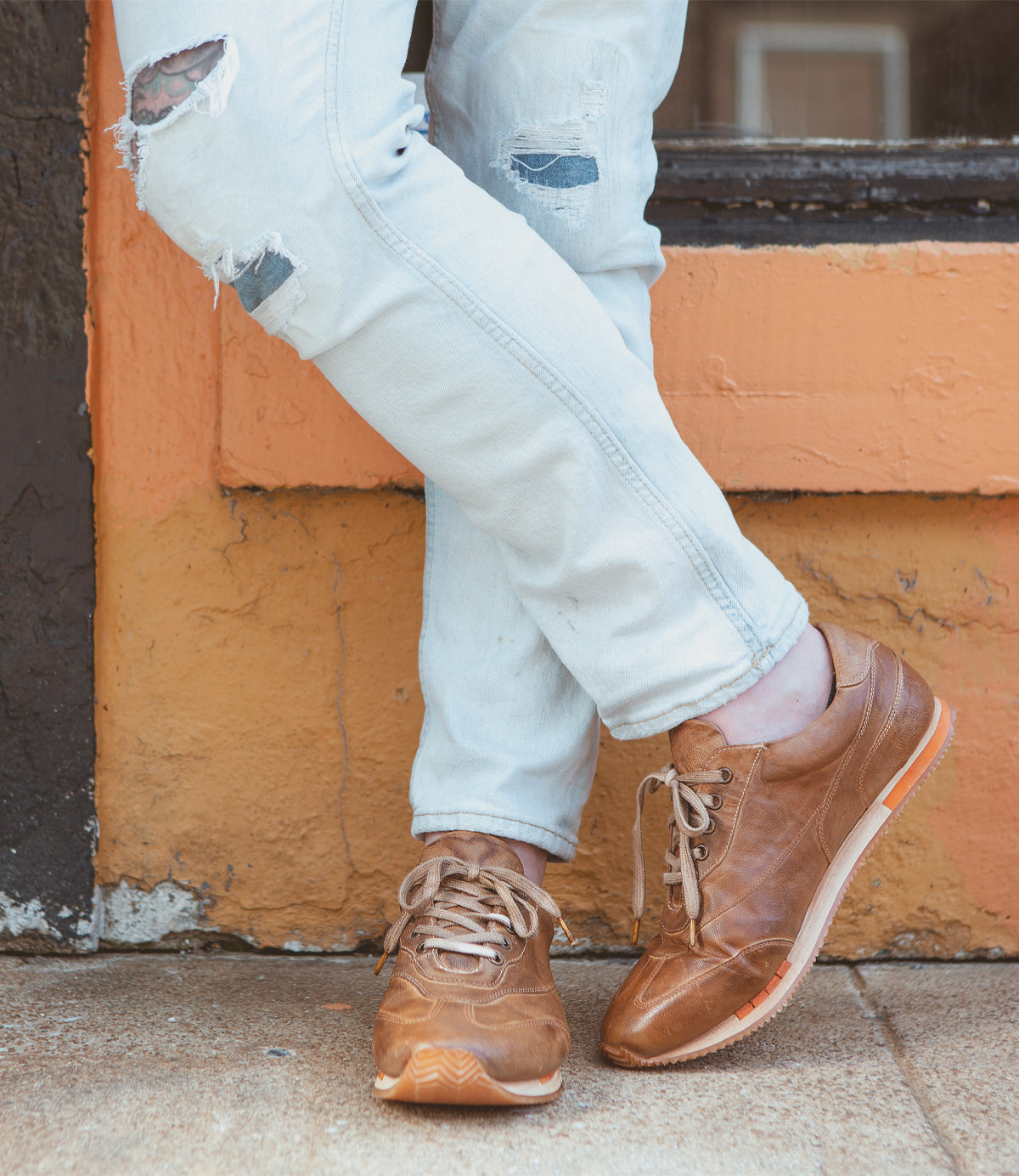 Person wearing ripped jeans and Roan Yann brown high-quality leather low profile sneakers leaning against an orange and yellow wall.