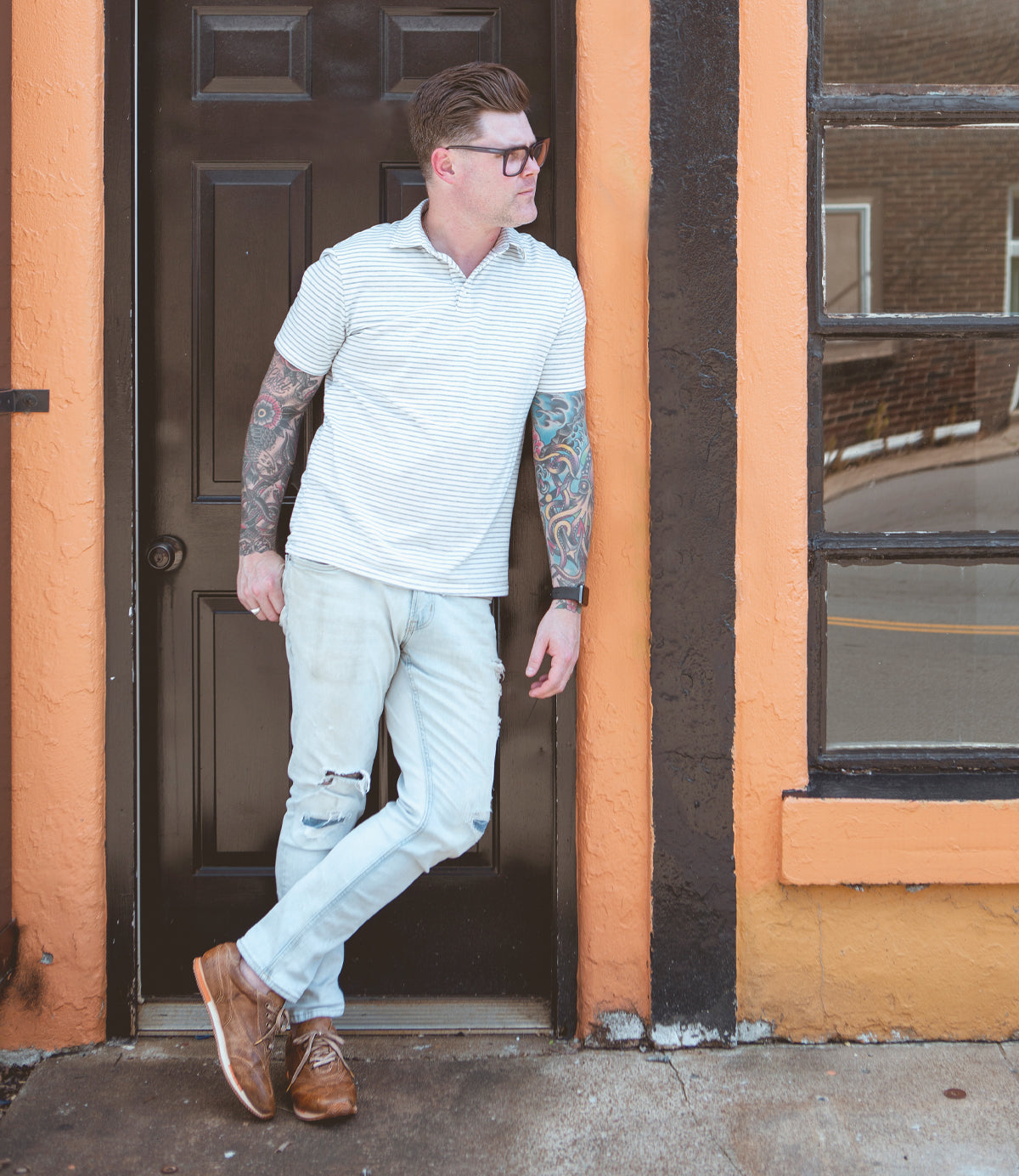 
                  
                    Man leaning against an orange door frame wearing sunglasses, a striped shirt, and light jeans with tattoos visible on his arms is sporting high-quality Roan Yann low-profile sneakers.
                  
                