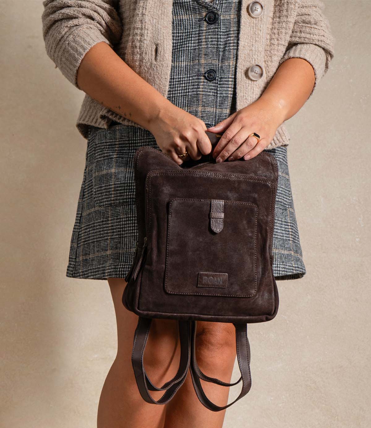 
                  
                    A woman in a plaid dress and textured jacket stands against a plain background, holding the Roan Yearling premium suede backpack with leather straps by its top handle.
                  
                