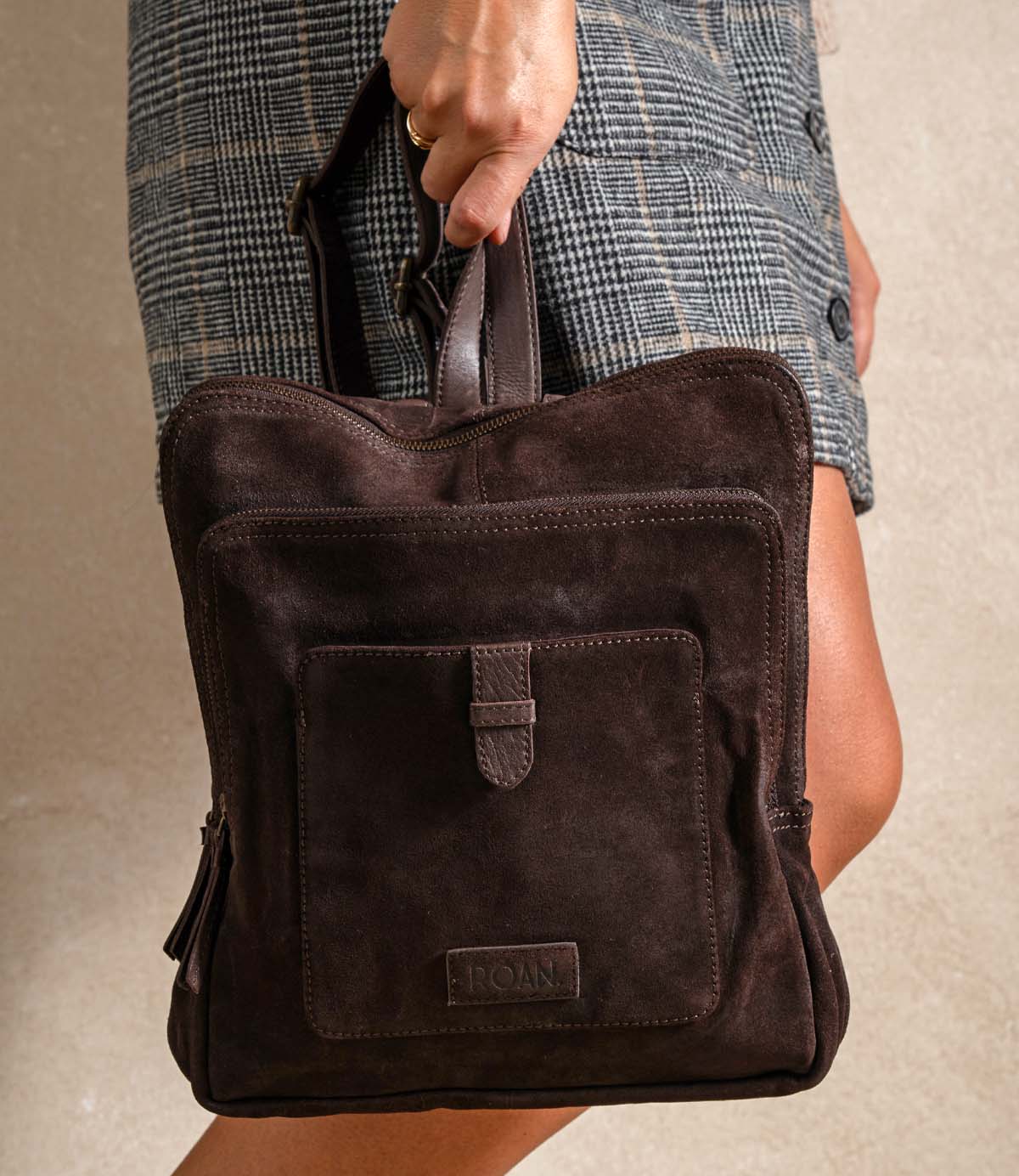 
                  
                    A person in a plaid skirt holds the Roan Yearling—a premium suede backpack with dual compartments, a front pocket, a leather handle, and stylish leather straps.
                  
                