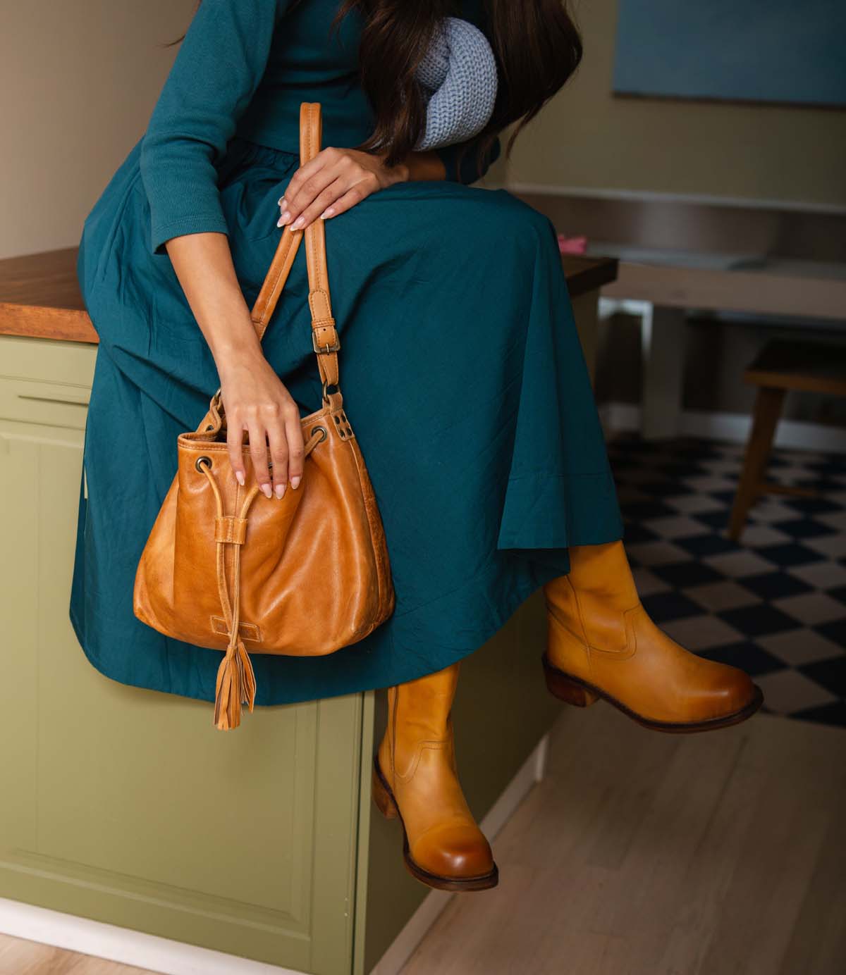 
                  
                    A person in a teal dress and tan leather boots holding a versatile, suede drawstring Zone bucket bag by Roan.
                  
                