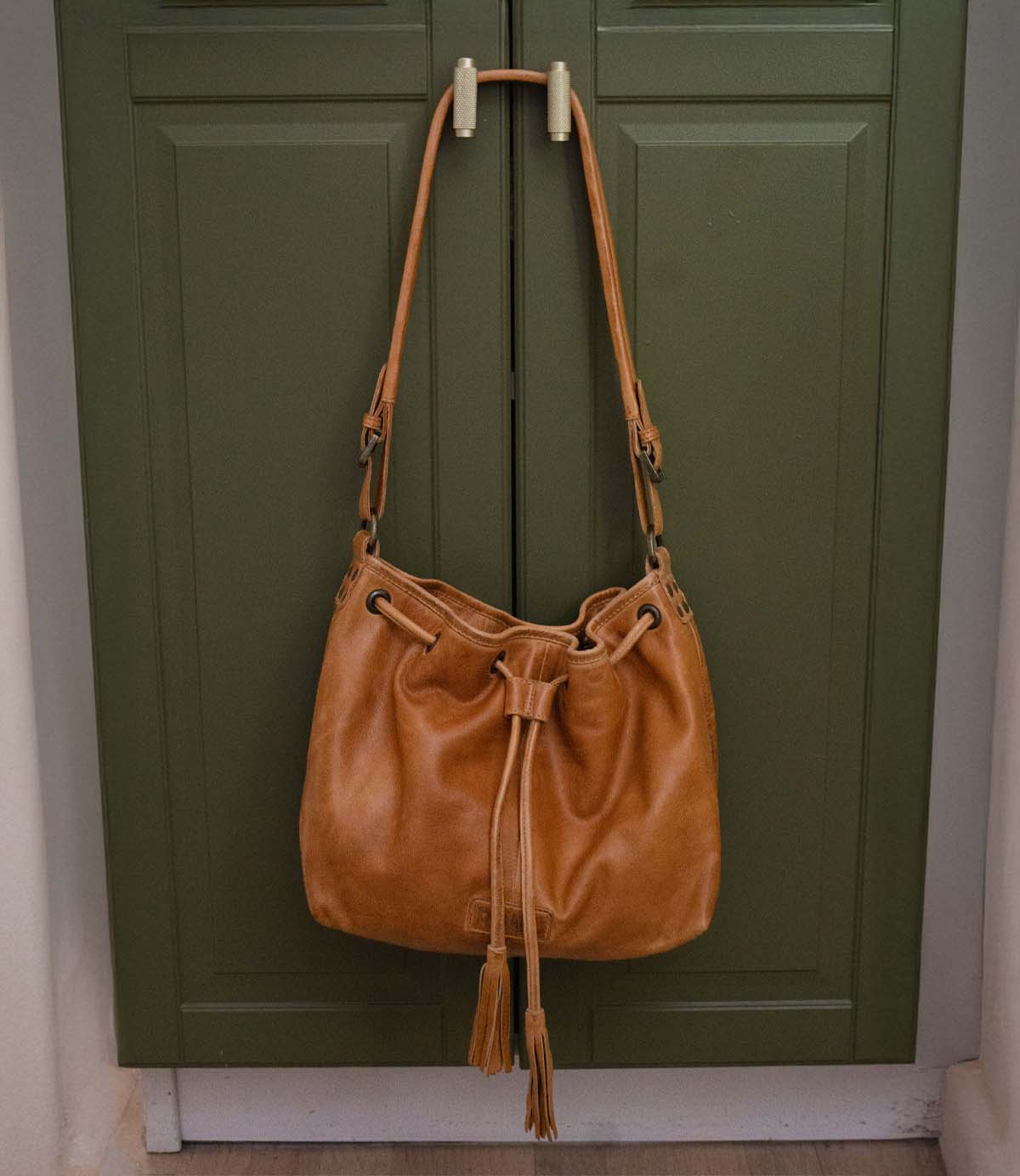 Roan Zone tan leather bucket bag with an adjustable strap, drawstring closure, and tassel detail, hanging on a cabinet door handle.